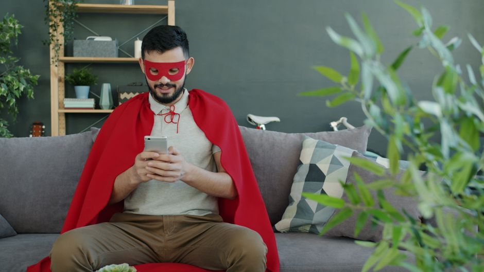 Young man in superhero costume using smartphone, relaxing on modern sofa indoors.